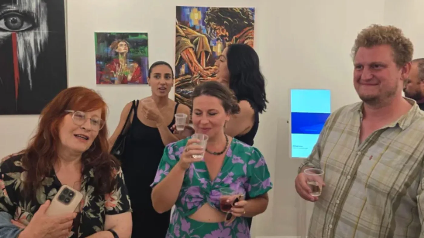 Guests holding drinks stand in a white gallery room hung with portraits and a large painted eye, with a small screen on the wall
