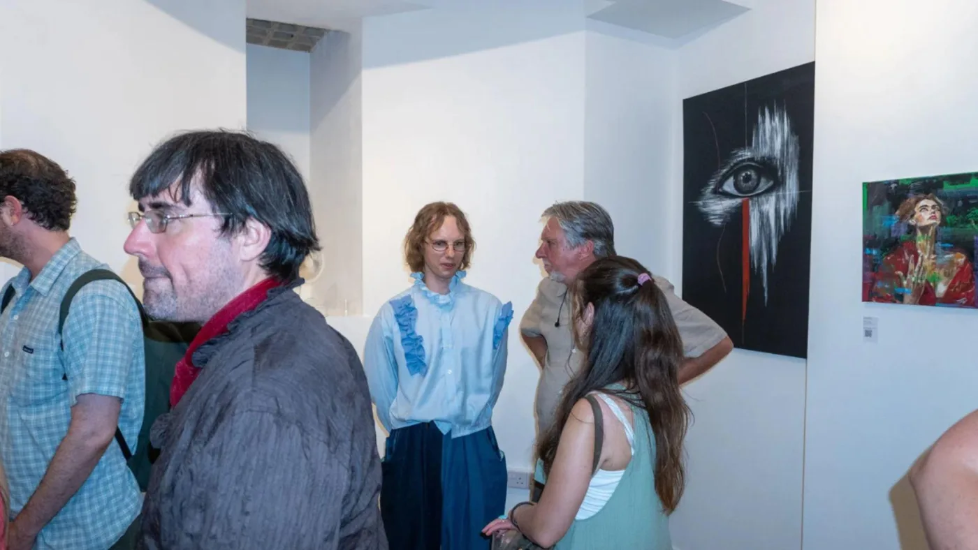 Visitors talk in a white-walled gallery beside a dark painting of a single eye and a colourful portrait