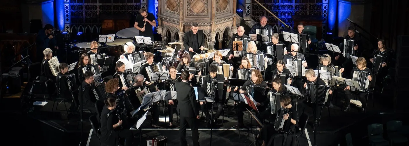 A large accordion orchestra seated in rows on a stage lit in blue, with a percussion section at the back and the conductor standing in front