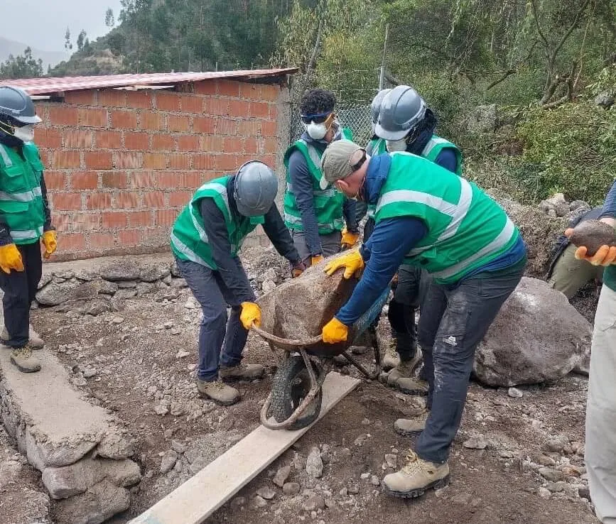Cadets in hard hats and green high-visibility vests moving a heavy rock in a wheelbarrow on the building site in Peru