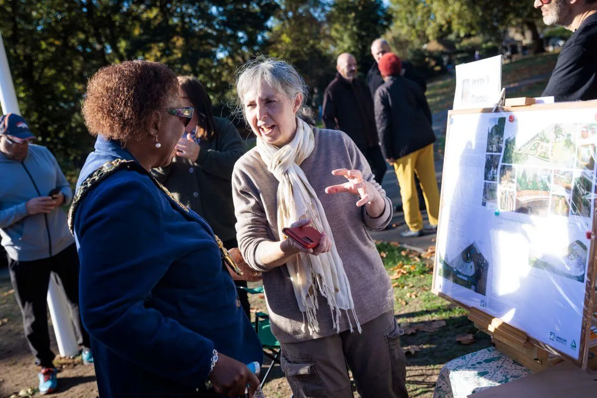 The Mayor of Hammersmith and Fulham, wearing her chain of office, talks with a volunteer beside a display board showing plans for the Jenny's Corner garden