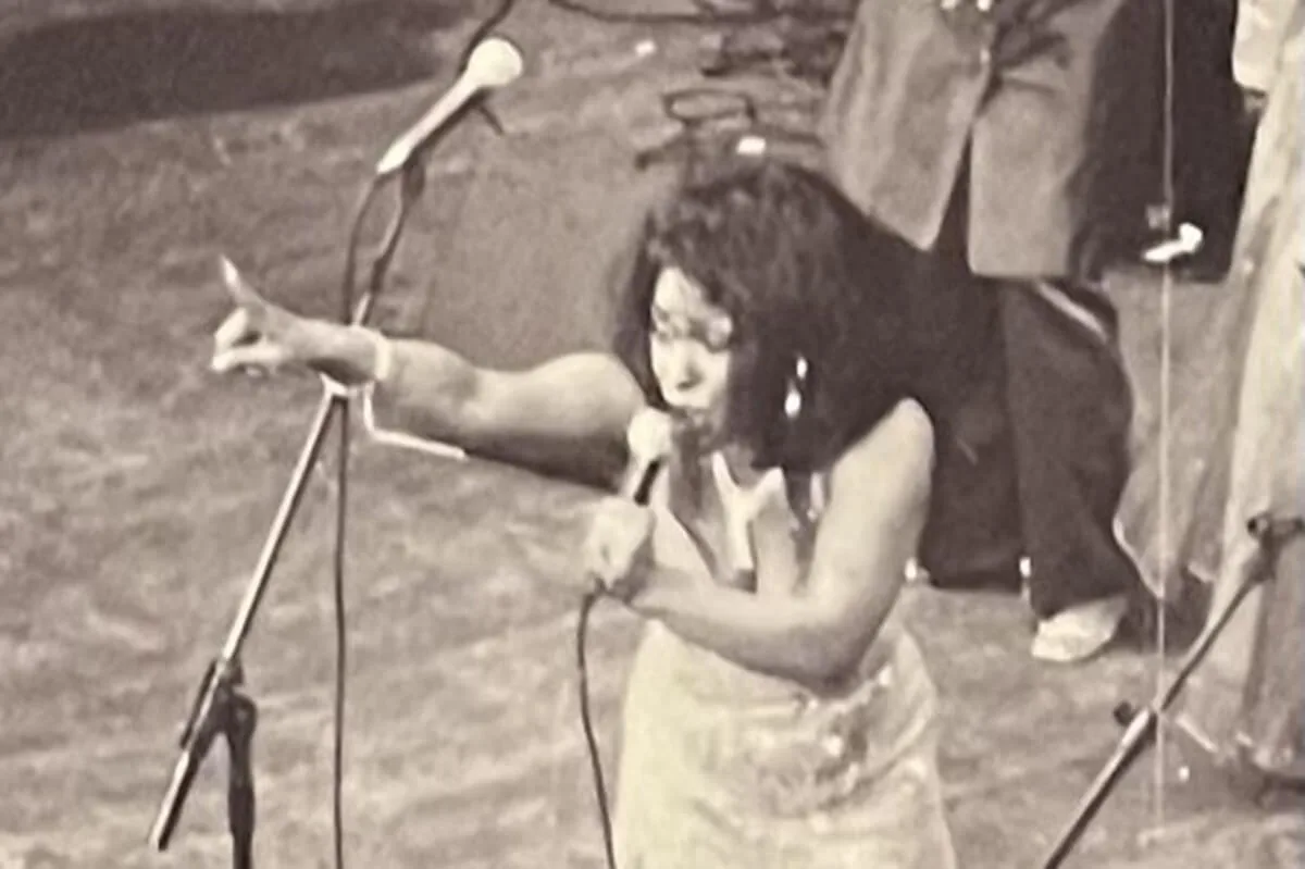 Louisa Mark singing into a hand-held microphone on stage, pointing out towards the audience