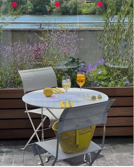 A small round white table with two drinks and a lemon, two folding chairs and planted borders on the terrace, with the river behind