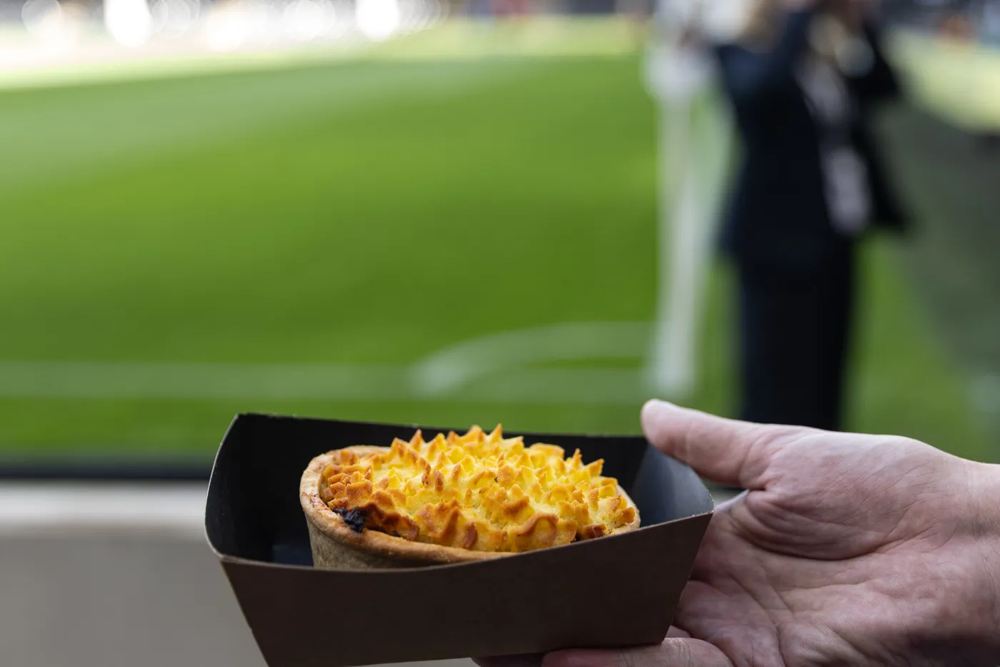 A hand holding a pie in a cardboard tray beside the pitch