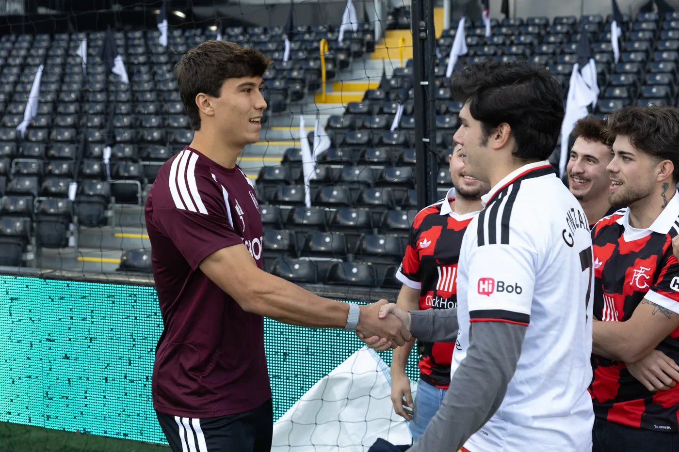 A member of the Fulham squad in a maroon training top shakes hands with fans from the group on the pitch at Craven Cottage