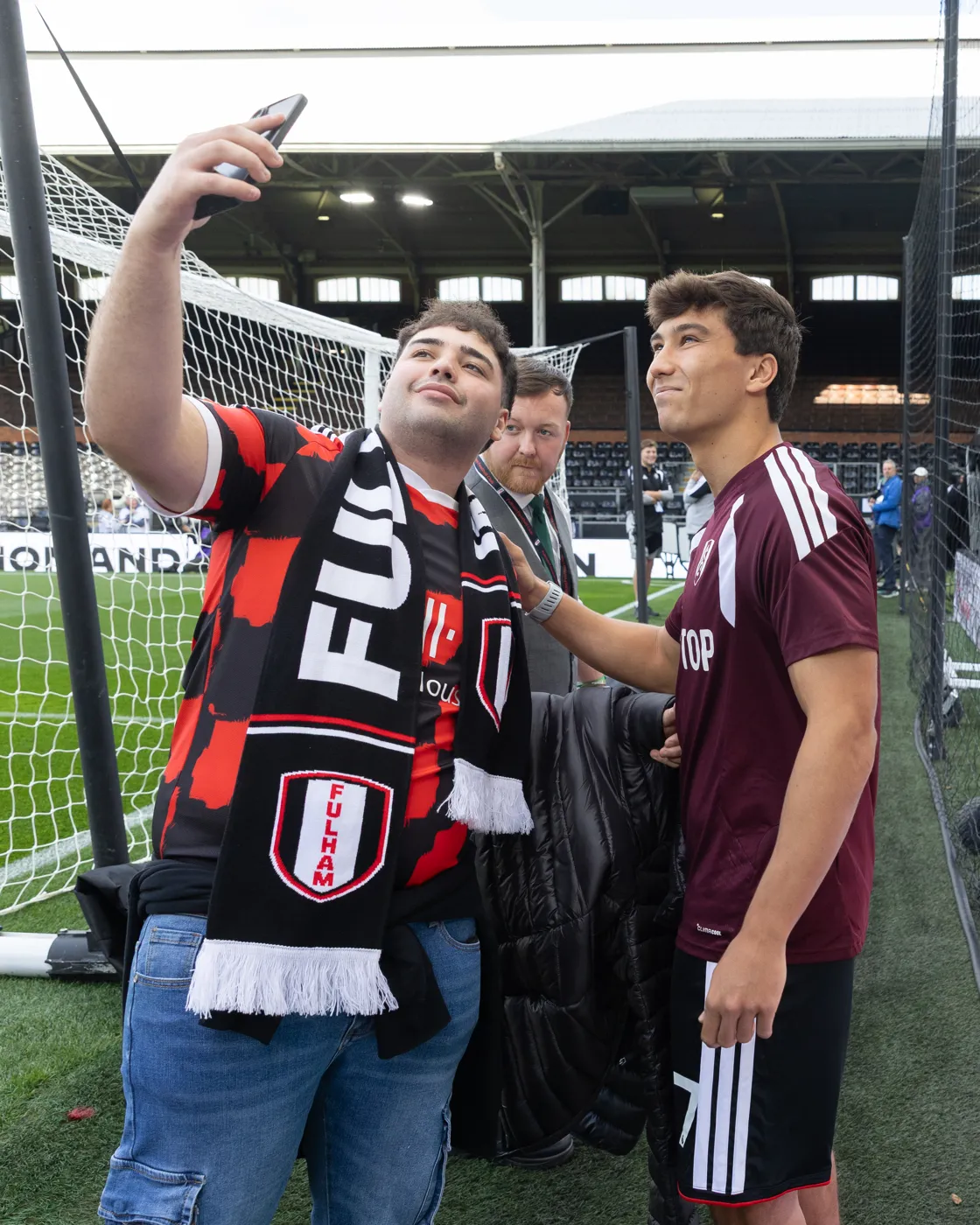 A fan in a Fulham scarf takes a selfie with a player in a maroon training top beside the goal