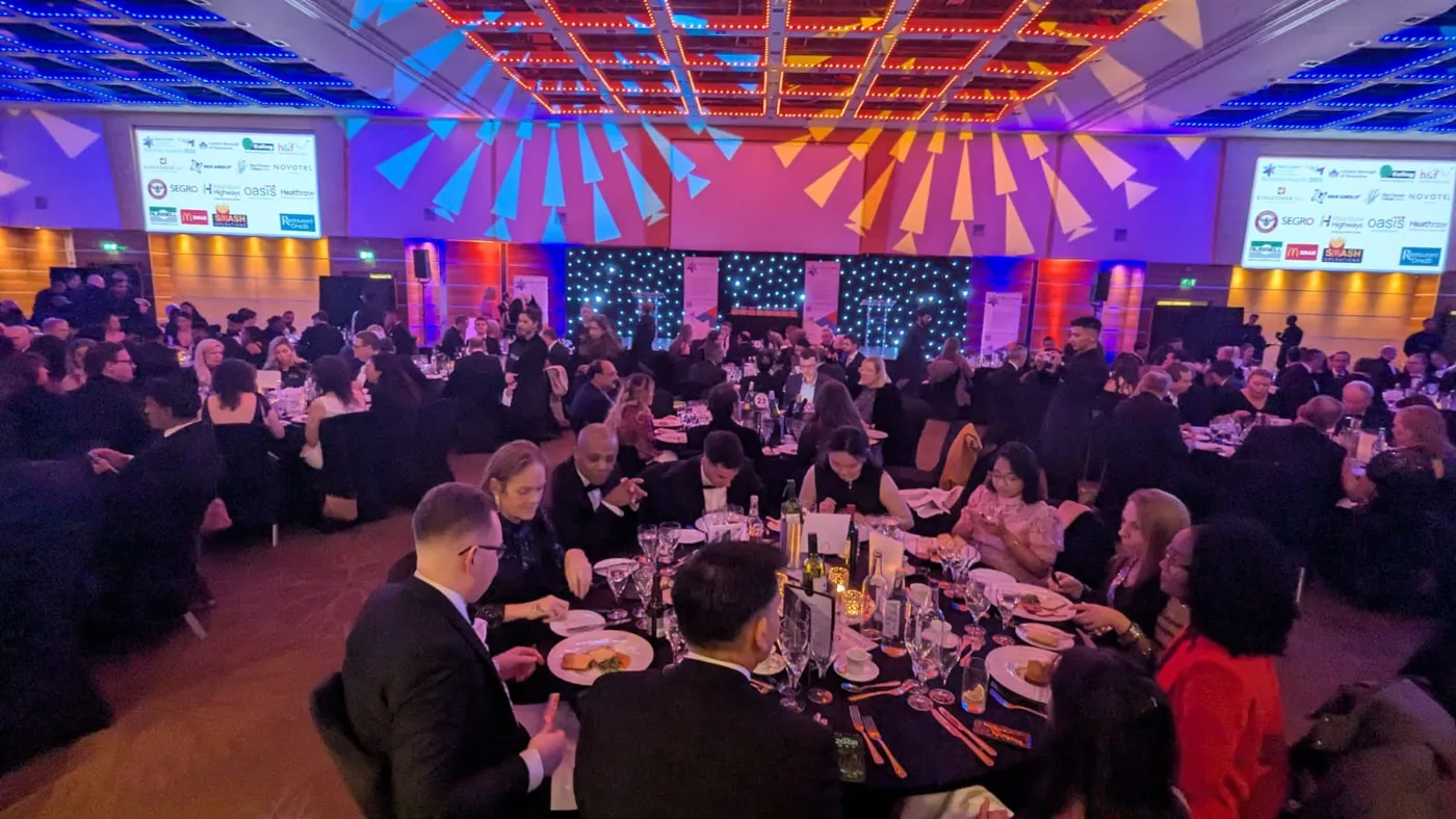 Guests in evening dress seated at round tables for dinner in a ballroom lit in red, blue and orange, with sponsor logos on screens
