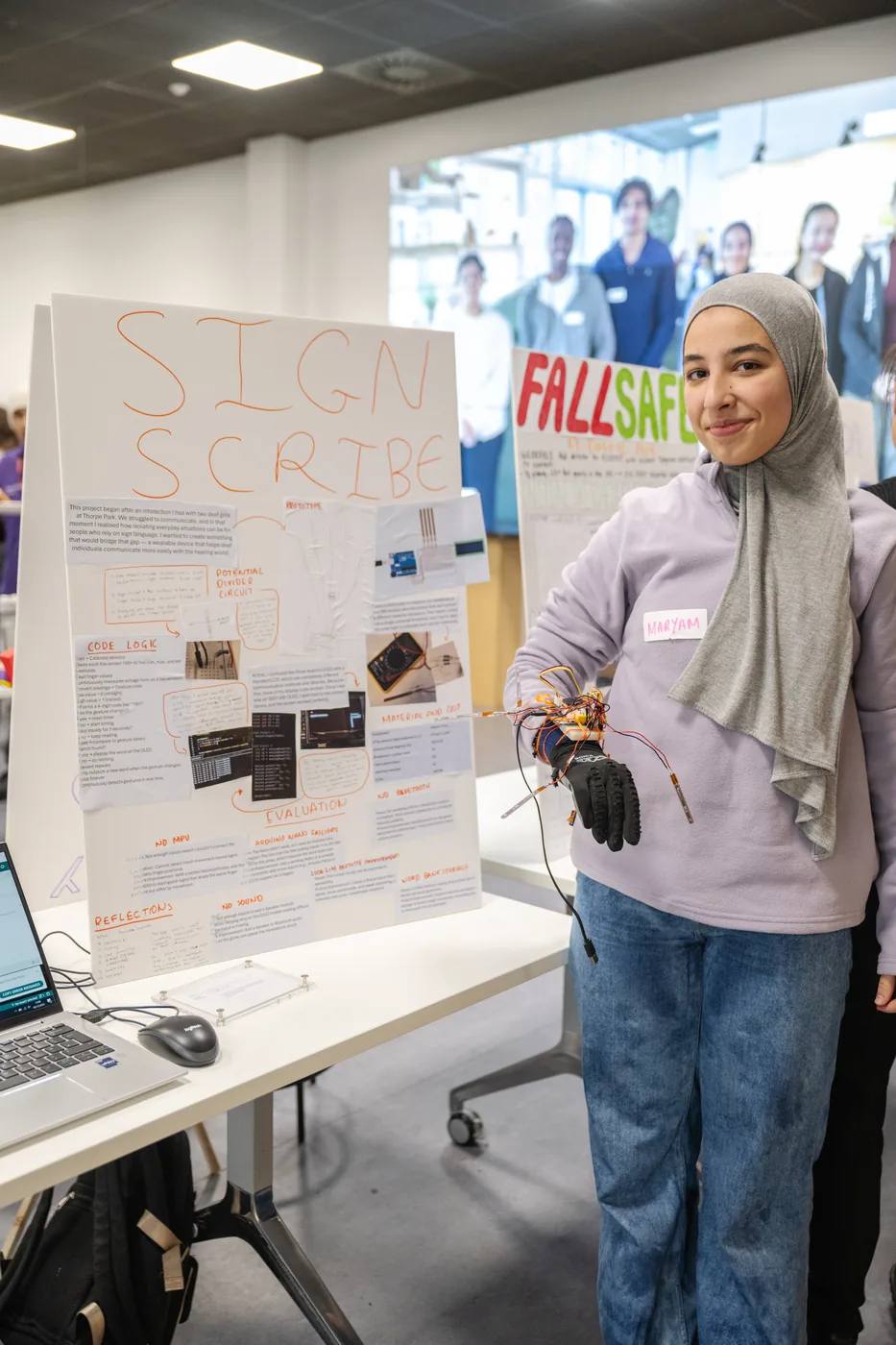 Maryam standing beside a large hand-drawn project board headed Sign Scribe, holding a wired glove, in a workshop space