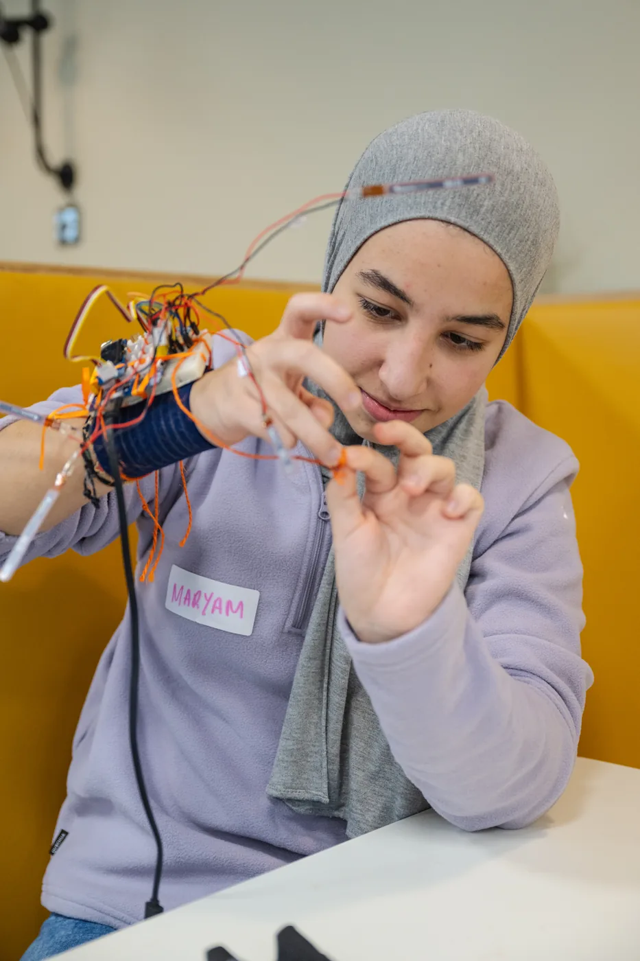 Maryam wearing the Sign Scribe glove, its orange wiring and circuit board visible along her forearm, flexing her fingers to test it