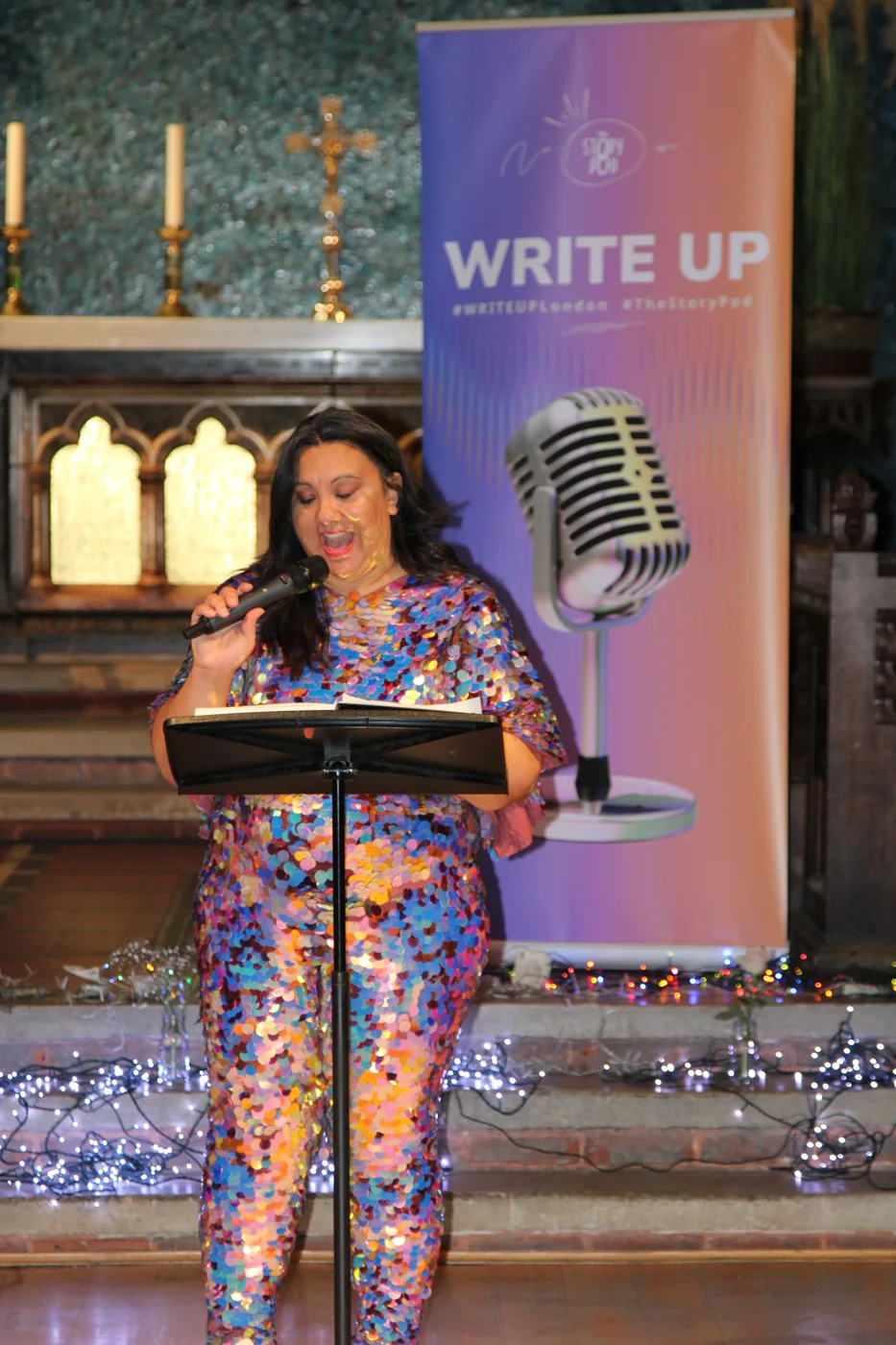 A woman in a sequinned outfit reading at a lectern beside a vintage-style microphone and a WRITE UP banner, in a church