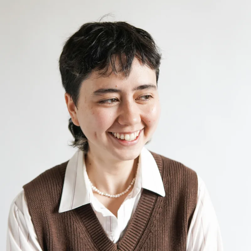 Vee Matsumari, with short dark hair, wearing a brown tank top over a white shirt and smiling